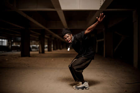 Emotional full length portrait of the dancing afro american man in the half squat with dreadlocks and dressed in black clothesの写真素材