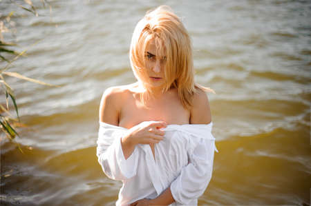 Nude portrait of beautiful and sexy blond woman in white shirt and with naked shoulders on the background of waterの写真素材