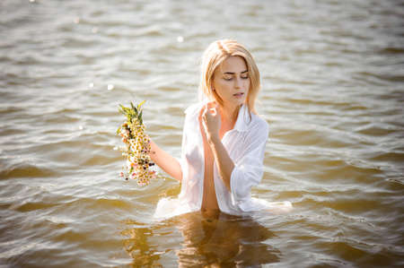 Nude portrait of a pretty blond girl in white wet shirt with wreath in hand, standing in waterの写真素材