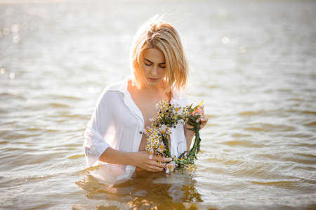 Nude portrait of a pretty blond girl in a white wet shirt with wreath in hands, standing in waterの写真素材