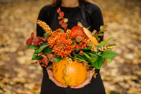 Woman in a black dress holding a beautiful pumpkin with lovely autumn flowers on the background of yellow leafsの写真素材