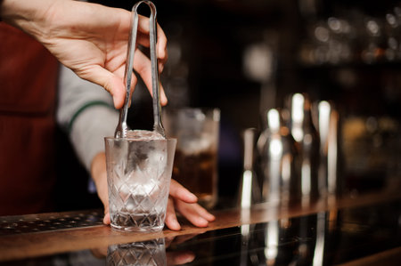Hand of bartender is putting an ice into a shot glass on the dark background