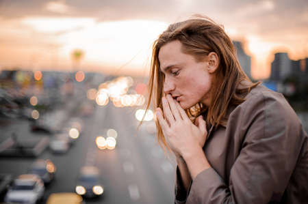 Side view of a young redhead man connecting palms near face and praying with closed eyes on the background of city and sunsetの写真素材