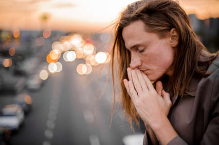 Side view of the young redhead man connecting palms near face and praying with closed eyes on the background of city and sunsetの写真素材