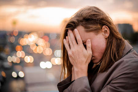 Redhead and longhaired young guy covering face with hands on the background of city and sunset. Side viewの写真素材