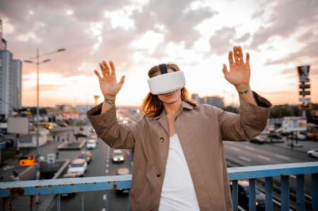 Young long-haired guy in brown jacket and in VR glasses on the blureed background of sunset and cityの写真素材