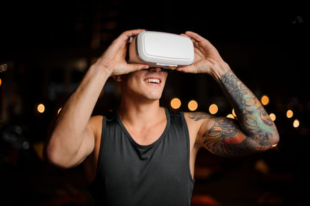 Muscular and tattooed smiling handsome man dressed in black shirt looks in night vision glasses on...