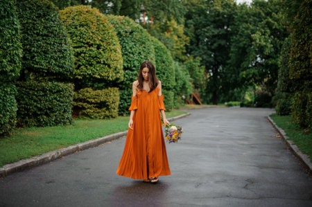 Attractive woman in long orange dress standing on the road with bouquet of flowers on the background of treesの写真素材