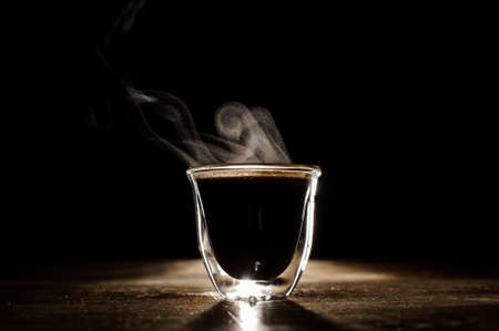 Minimalistic photo of glass mug with coffee and steam from it located on the wooden tableの写真素材