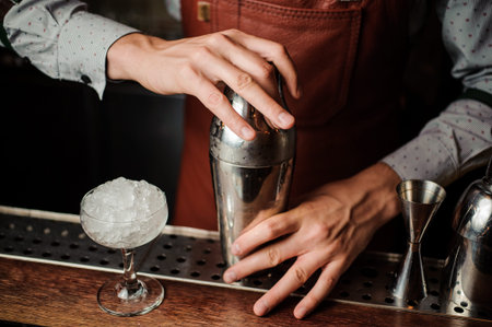 Barman in brown apron holding a shaker in hands behind the bar counter on the indoors backgroundの写真素材