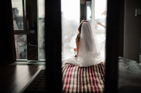 Back view of beautiful bride in wedding veil sitting on the bed against the background of large windowの写真素材