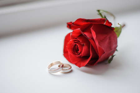 Two golden wedding rings near the red rosebud arranged on the white background of window sillの写真素材