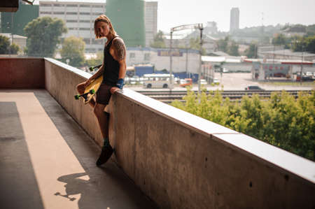 Handsome long-haired guy with tattoo sitting on the parapet and holding a longboard on the background of cityの写真素材