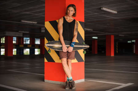 Portrait of the handsome young redhead man leaning on the pillar and holding a longboardの写真素材