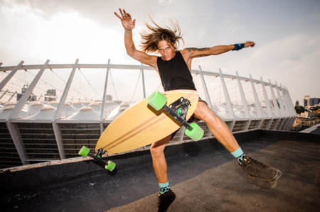 Young long-haired and redhead guy jumping high on a longboard against the outdoors background of sky and roofの写真素材