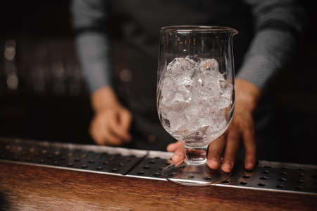 Male barman holding a large glass filled with ice cubes on the wooden bar counterの写真素材
