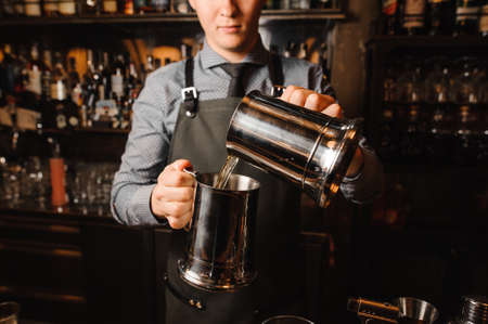 Young bartender in a grey shirt and apron pouring a clear alcoholic drink on the bar counterの写真素材