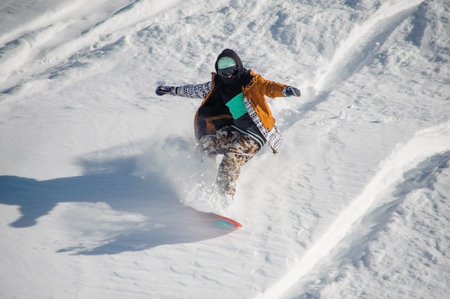 Active snowboarder in colorful and stylish sportswear riding with snowboard down the powder snow...