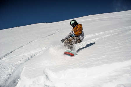 Snowboarder in sportswear riding on the powder mountain slope against the scenic background of bright blue skyの写真素材