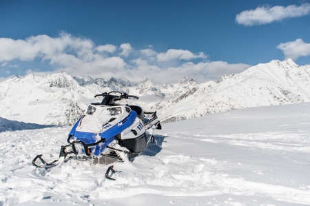 Modern blue snow vehicle against the scenic background of high snow mountains peaks and cloudy skyの写真素材