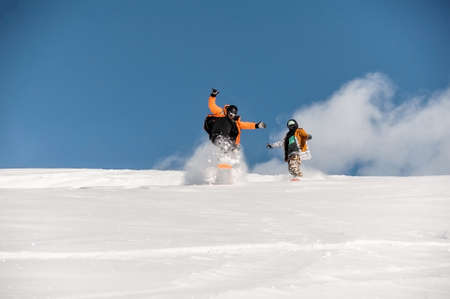 Couple of snowboarders in sportswear riding on the powder mountain slope against the scenic background of bright blue skyの写真素材