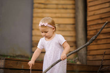 Lovely little blonde girl dressed in cute pink dress playing outdoors with a big stickの写真素材