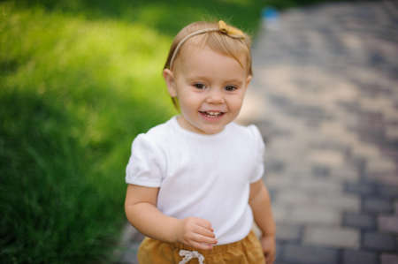 Smiling little blonde girl with cute headband walking outdoors near the fresh green grass lawnの写真素材