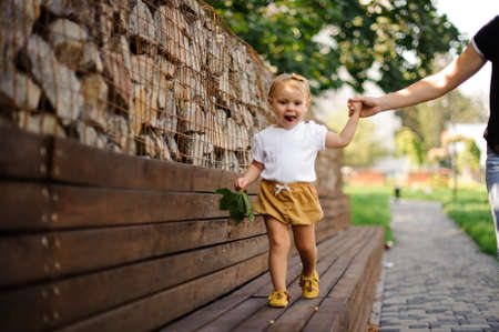 Pretty little blonde girl in cute clothing walking on the bench with help of mother s handの写真素材