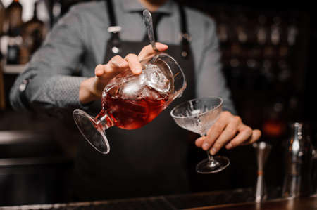 Barman pouring fresh alcoholic drink into the cocktail glass against the blurred background of bottlesの写真素材