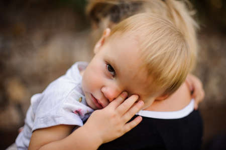 Little blonde child dressed in a white shirt lying on mom's shoulder and wiping eyesの写真素材