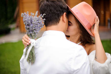 Groom in white shirt hugging bride in white dress and pink hat with bouquet of flowers on the outdoors backgroundの写真素材