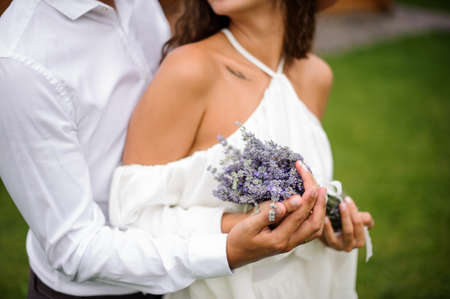 Groom in white shirt hugging bride in white dress with bouquet of lavender on the blurred outdoors background of green grassの写真素材