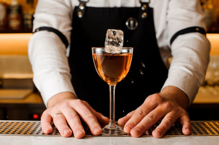 Barman in a white shirt and apron holding a glass with alcoholic drink and ice cube on the bar...