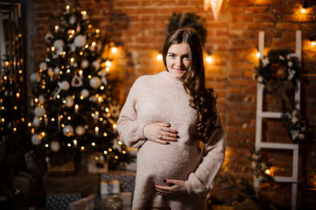 Beautiful pregnant woman with long hair on the background of Christmas tree and garland lightsの写真素材