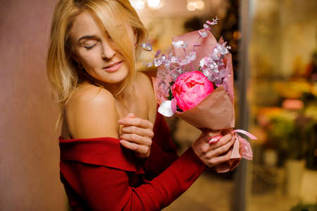Young blonde woman with closed eyes holding a pretty bouquet of pink flowers wrapped in a paperの写真素材