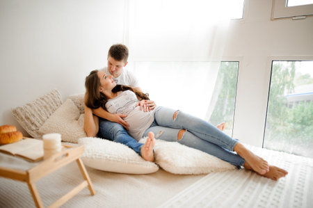 Portrait of man with beautiful pregnant woman lying on the large bed in a white room. Pregnancy...