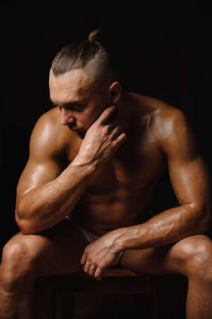 Hot athletic and muscular young man with stylish haircut sitting and looking down on the black background of studioの写真素材