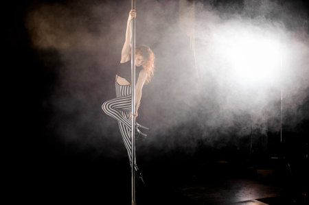 Young and sexy red haired woman exercises pole dance in the smoke on the dark background of studio
