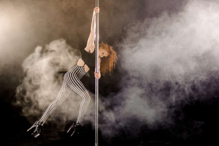 Red haired sexy pole dance girl exercises and poses on the pylon in the smoke on the black backgroundの写真素材