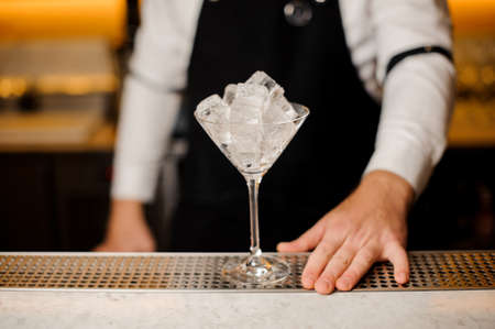Elegant cocktail glass filled with ice cubes on the bar counter against the bartender dressed in apron and white shirtの写真素材