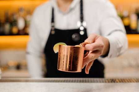Bartender in white shirt and apron holding a cup filled with ginger drink and decorated with a slice of limeの写真素材