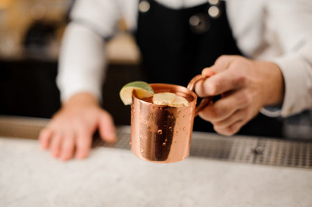 Bartender in white shirt and apron holding a cup filled with a fresh ginger drink and decorated with a slice of limeの写真素材