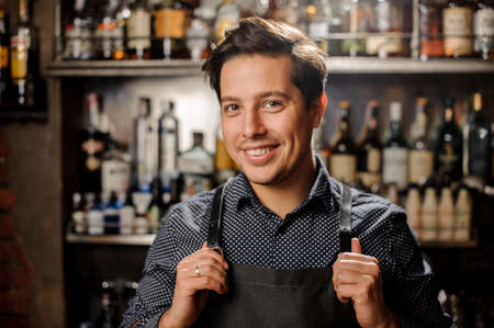 Handsome and young smiling brown haired barman on the background of shelves with bottles of alcoholの写真素材
