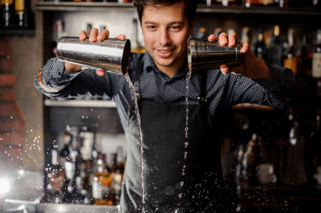 Young smiling barman pouring alcoholic drink from his shakers on the background of bar counterの写真素材