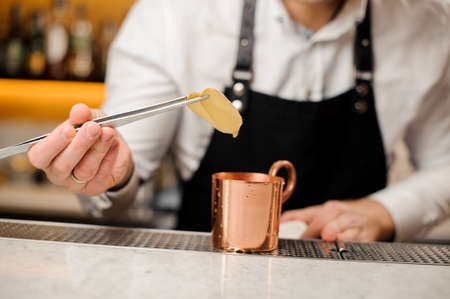 Bartender in a white shirt and black apron adding two slices of ginger into a metal cupの写真素材