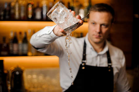Barman dressed in a white shirt pouring water from a glass filled with ice cubesの写真素材