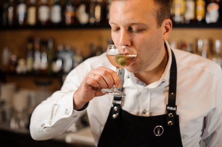 Young barman dressed in a white shirt drinking alcoholic cocktail with green olives on the...