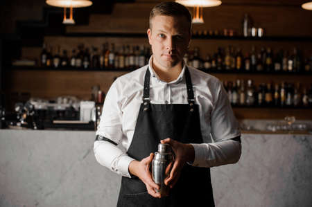 Bartender in white shirt and apron holding a shaker against the blurred indoors background of the bar counterの写真素材