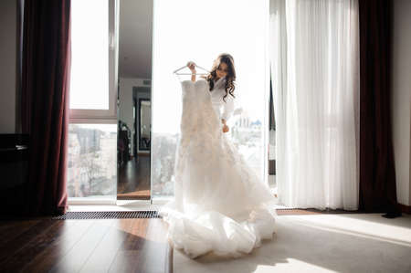 Pretty brunette bride holding delicate white wedding dress on a hanger on the background of view from the windowの写真素材