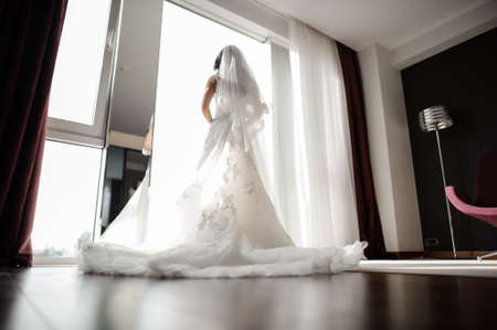 Elegant romantic brunette bride in white wedding dress and silk veil looking through the windowの写真素材